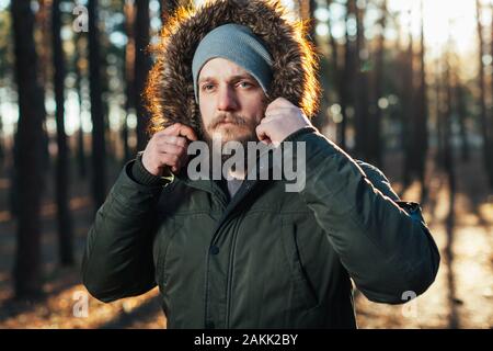 Porträt, Nahaufnahme der Jungen stilvoll ernster Mann mit Bart in rgreen Winter Jacke mit Kapuze und Fell auf dem Kopf bekleidet steht gegen die Rückseite Stockfoto