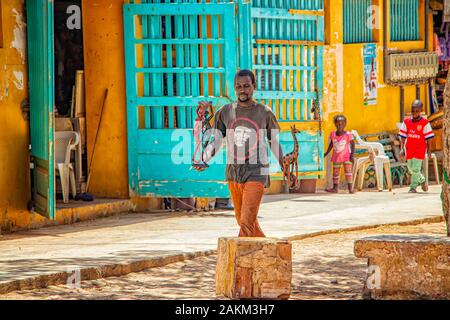 Insel Goree, Senegal - 22. April 2019: unbekannter Mann Spaziergang entlang einem sandigen Straße in einer kleinen Stadt und eine aus Holz geschnitzte Figuren tragen. Stockfoto