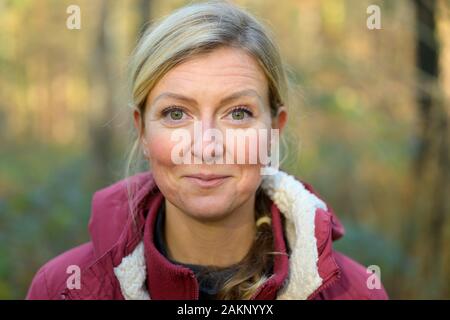 Nach blonde Frau in den Vierzigern, trägt warme rote Jacke lächelnd und mit Blick auf die Kamera, während er draußen im Wald. Büste Front-buchmontage Stockfoto