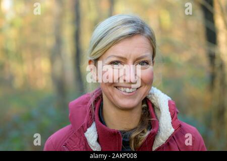 Nach blonde Frau in den Vierzigern, trägt warme rote Jacke lächelnd und mit Blick auf die Kamera, während er draußen im Wald. Büste Front-buchmontage Stockfoto