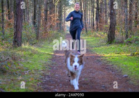 Lächelnde Frau Joggen im Freien mit ihrem Hund läuft auf einem unbefestigten Weg in den Wald oder Park Stockfoto