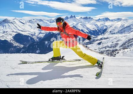 Skifahrerin, die sich an einem sonnigen Wintertag mit Les Aiguil auf Skiern mit lustiger Pose hoch oben auf einer Skipiste in der Skidomäne Les Sybelles, Frankreich, streckt Stockfoto