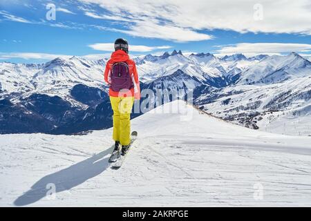 Skifahrerin mit farbenfroher Kleidung, die auf die Gipfel von Les Aiguilles d'Arves blickt, von einer Skipiste hoch über der Skidomäne Les Sybelles, in Frankreich Stockfoto