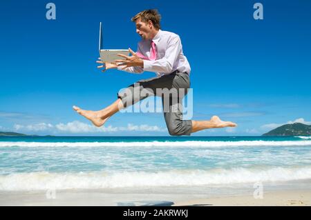 Aufgeregt Geschäftsmanns, der auf seinem Laptop am Ufer eines leeren tropical beach springen Stockfoto