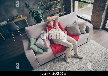 Oben über dem hohen Blickwinkel auf sie schöne müde kranke, kranke, grauhaarige Oma, die rotes Kostüm trägt, das auf Divan auf dem industriellen Ziegelboden liegt Stockfoto