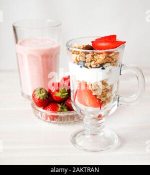 Erdbeer-Milchshake aus Glas, Erdbeeren in Cristal Schüssel, Granola und Joghurt Dessert mit Blaubeeren in hoher Glasmücke auf weißem Vintage-Holzbac Stockfoto