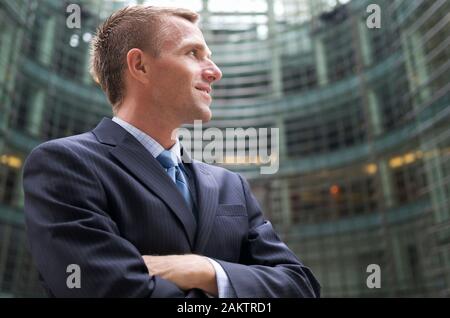 Selbstbewusster Geschäftsmann, der vor einem Bürogebäude steht und in dunklem Nadelstreifenanzug lächelt Stockfoto