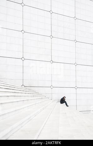 Entfernte Figur eines Mannes, der mit dem Kopf in den Händen huscht und auf einer großen weißen Treppe sehr klein aussieht Stockfoto