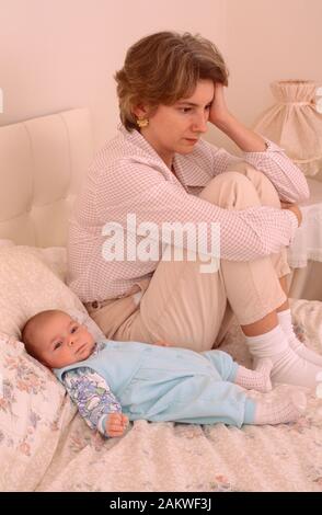 Junge Mutter mit einem Baby, das an einer postnatalen Depression leidet, traurig aussieht und auf einem Bett sitzt Stockfoto