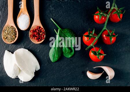 Blick auf italienische Zutaten: Frische Kirschtomaten, Mozzarella, Gewürztablespoon, Knoblauch, Spinat. Gesunde mediterrane Ernährung. Dunkles Backgro-Licht Stockfoto