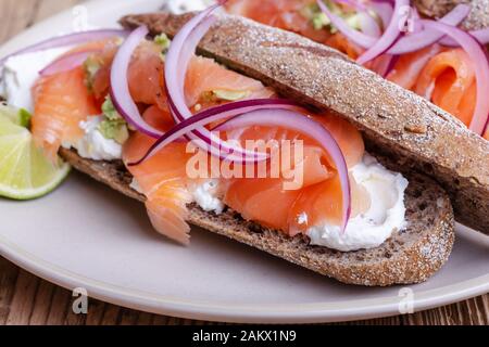 Gehärteter Lachs ganze Weizenbaguette Sandwiches, Brot mit Quark, geräucherter Fisch, rote Zwiebel, Avocado auf rustikalem Tisch in der Nähe Stockfoto