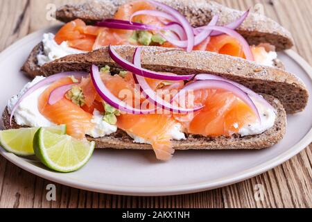 Gehärteter Lachs ganze Weizenbaguette Sandwiches, Brot mit Quark, geräucherter Fisch, rote Zwiebel, Avocado auf rustikalem Tisch in der Nähe Stockfoto