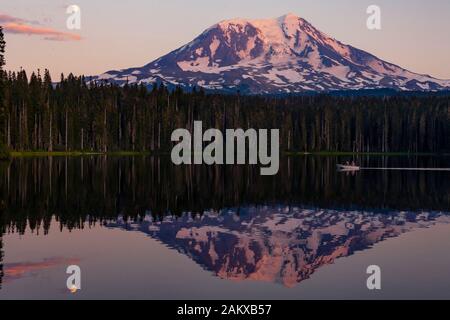 Der Mond ragt über den Mount Adams auf und eine Spiegelung wird im ruhigen Takhlakh Lake bei Sonnenuntergang gesehen. Familienboater genießen Sommerwetter in der Ferne. Stockfoto