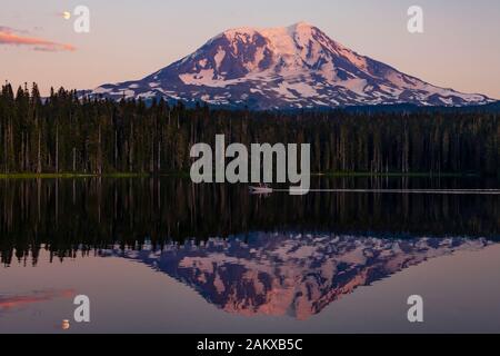 Der Mond ragt über den Mount Adams auf und eine Spiegelung wird im ruhigen Takhlakh Lake bei Sonnenuntergang gesehen. Familienboater genießen Sommerwetter in der Ferne. Stockfoto