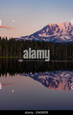 Der Mond ragt über den Mount Adams auf und eine Spiegelung wird im ruhigen Takhlakh Lake bei Sonnenuntergang gesehen. Familienboater genießen Sommerwetter in der Ferne. Stockfoto