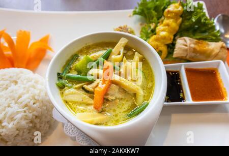 Eine Schüssel mit traditionellen thailändischen grüne Chicken Curry mit Reis, ein Satay, eine Frühlingsrolle und taucht in eine Porzellanschüssel auf einem weißen Teller serviert Stockfoto