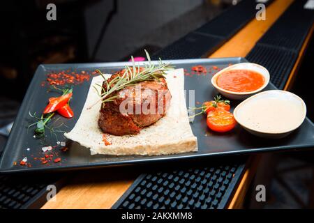 Ein saftiges gegrilltes Steak mit zwei Soßen sitzt auf einem schwarzen Teller. Warme Fleischgerichte, Oberteil mit Rosmarinsprchen dekoriert. Stockfoto