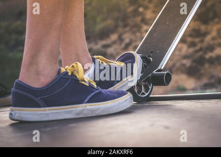 Nahaufnahme der Füße eines Teenagers auf einem Roller, der bereit ist, eine Fahrt über die Halfpipe zu beginnen. Skater-Startsprünge und Tricks im Skatepark. Lassen Sie uns genießen Stockfoto