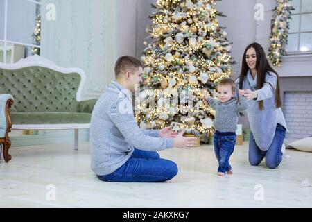 Glückliches junges Paar und ihr Baby-Sohn in der Nähe des Weihnachtsbaums zu Hause. Mutter unterstützt kleinen Jungen, während er erste Schritte in Richtung Vater macht. Stockfoto