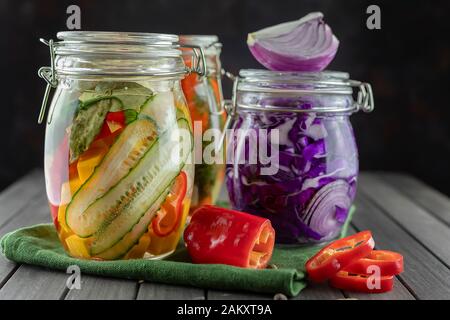 Drei Glasgefäße aus fermentiertem Rotkohl, Gurken, Chili, Gemüse auf dunklem Grund. Mit textilgrünem Material. Gärung ist eine Quelle Stockfoto