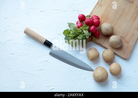 Frische Radieschen und Kartoffeln mit großem Küchenmesser auf weißem Grund und alter Holzschneideplatte. Kopierraum Stockfoto