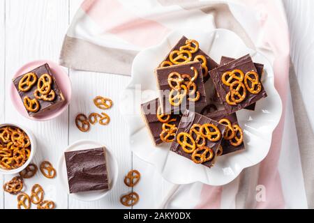 Nahaufnahme von Schokoladen-Peanut-Butterbars mit Brezel-Crackern auf einem Kuchenständer auf einem weißen Holztisch, Blick von oben, flacher Lay Stockfoto