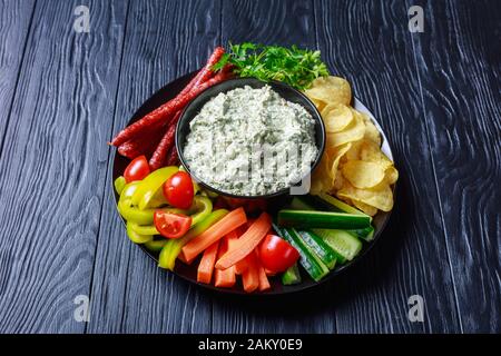 Nahaufnahme von Cremigem Spinat in einer Schüssel mit Würstchen, Karotten, Gurken, süßen Pfefferstöcken, Kartoffelchips und frischer Petersilie auf einem Teller, Blick von a Stockfoto