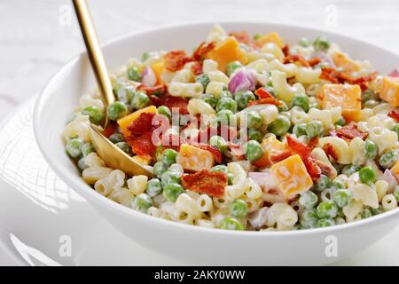 Nahaufnahme von Makkaroni-Salat mit grünem Erbsen, gebratenem knusprigem Speck, Cheddar-Käse und roter Zwiebel in einer weißen Schüssel mit goldenem Löffel, horizontaler Ansicht Stockfoto