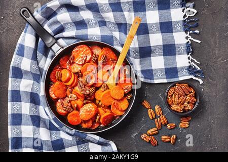 Kandierte Süßkartoffeln mit braunem Zucker, Ahorn-Sirup, Orangensaft und Pekannüssen in einem Skillet auf einem Betontisch mit Küchentuch, Ansicht von oben Stockfoto