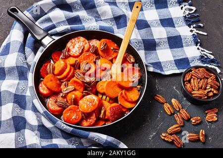 Kandierte Süßkartoffeln mit braunem Zucker, Ahorn-Sirup, Orangensaft und Pekannüssen in einem Skillet auf einem Betontisch mit Küchentuch, Ansicht von oben Stockfoto