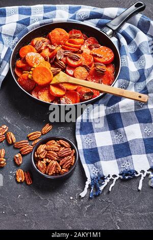 Kandierte Süßkartoffeln mit braunem Zucker, Ahorn-Sirup, Orangensaft und Pekannüssen in einem Skillet auf einem Betontisch mit Küchenhandtuch, vertikaler Ansicht f Stockfoto