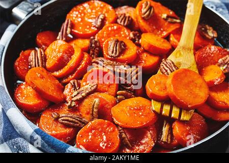 Kandierte Süßkartoffeln mit braunem Zucker, Ahorn-Sirup, Orangensaft und Pekannüssen in einem Skillet mit Holzspachtel, Blick von oben, Nahaufnahme, Makro Stockfoto