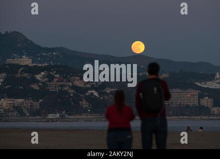 Die Menschen sehen den Vollmond (wie in diesem Monat als "Wolf moon" bekannt), als es über den Horizont am Strand steigt während der ersten partielle Mondfinsternis des Jahres. In diesem Jahr 2020 haben sechs Finsternisse, einschließlich einer totalen Sonnenfinsternis. Stockfoto