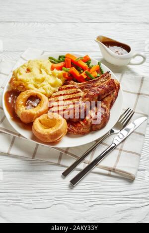 Gegrillte Schweinekoteletts auf Knochen, Kartoffelbrei, grünen Bohnen, Karotte, Yorkshire Puddings und Zwiebelgravy auf einem weißen Teller auf einem Holztisch Stockfoto