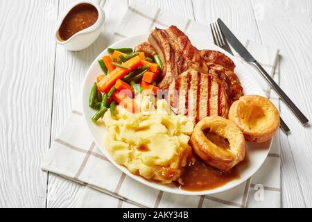 Nahaufnahme gegrillter Schweinekoteletts auf Knochen, Kartoffelbrei, grünen Bohnen, Karotte, Yorkshire Puddings und Zwiebelgravy auf einem weißen Teller auf einem Holztisch Stockfoto