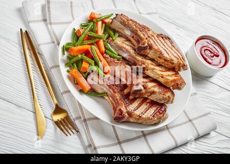 Nahaufnahme von Gegrillten Schweinekoteletts an Knochen, serviert mit grüner Bohne und Karottensalat auf einer weißen Platte auf einem Holztisch mit goldener Gabel und Messer, Stockfoto