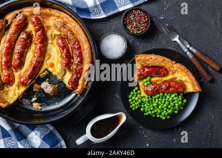 Nahaufnahme von "Toad in the Hole", "Wurst Toad", "Würstchen in Yorkshire Pudding" in schwarzer Keramikbackform und eine Portion auf einem Teller mit grünen Erbsen Stockfoto