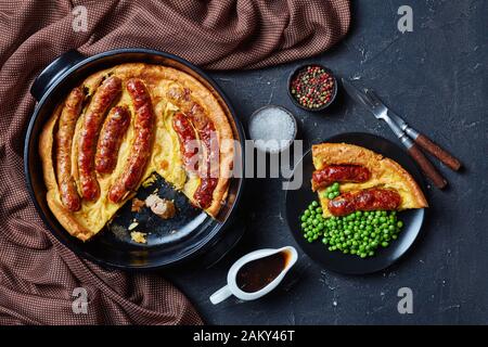 Kröte im Loch, Wurst Toad, Würstchen in Yorkshire Pudding-Teig in schwarzer Keramikschale und eine Portion auf einem Teller mit grünen Erbsen und Zwiebeln g Stockfoto