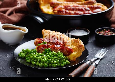 Nahaufnahme einer Portion Toad in the Hole, Wurst Toad, Würstchen in Yorkshire Pudding-Batter auf einem schwarzen Teller mit grünen Erbsen und Zwiebelgras, Stockfoto