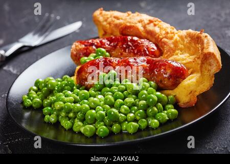 Nahaufnahme einer Portion Toad in the Hole, Wurst Toad, Würstchen in Yorkshire Pudding-Batter auf einem schwarzen Teller mit grünen Erbsen und Zwiebelgras, Stockfoto