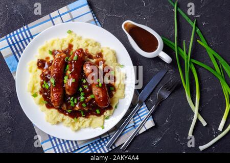 Klassische Banger und Mash mit Zwiebelgravy bestreut mit Frühlingszwiebel auf einem Teller auf einem Betontisch, englische Küche, flaches Lay Stockfoto