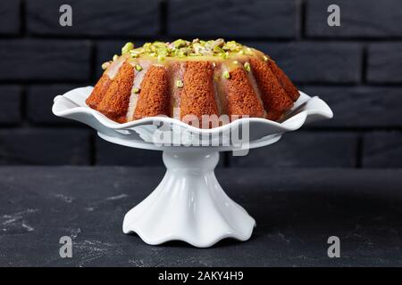 Köstlicher feuchter Kürbis-Karotten-Bundkuchen mit orangefarbener Glasur, mit Pistazien auf einem weißen Keramikständer mit einer Ziegelwand am Hintergrund bestreut, c Stockfoto