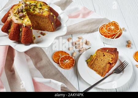 Köstlicher feuchter Kürbis-Karotten-Biskuit mit orangefarbener Glasur, mit Pistazien auf Keramikständer und einer Scheibe auf einem Teller auf einem weißen Holztabl bestreut Stockfoto