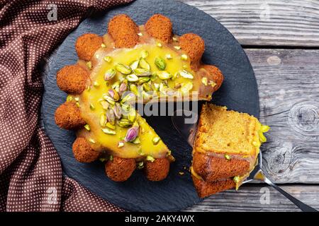 Kürbis-Karotten-Biskuitkuchen mit orangefarbener Glasur, mit Pistazien auf schwarzem Teller auf rustikalem Holztisch, Flachlay, Makro bestreut Stockfoto