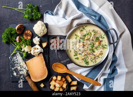 Asiago Geröstete Knoblauchkaulblumensuppe in einem Stocktopf mit Zutaten und Croutons auf einem Betontisch, horizontaler Blick von oben, flacher Lay Stockfoto