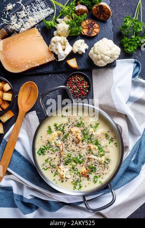 Nahaufnahme von Asiago Gerösteter Knoblauchkaulblumensuppe in einem Vorratsbehälter mit Zutaten und Croutons auf einem Betontisch, vertikale Ansicht von oben Stockfoto