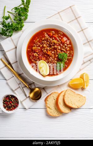 Linsensuppe mit Gemüse und Zitronenkeil in einer weißen Schüssel auf einem Holztisch mit goldenem Löffel und Toast, vertikale Ansicht von oben Stockfoto