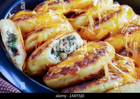 Nahaufnahme hausgemachter Kartoffelkuchen mit Pilzen und Dillfüllungen mit geriebenem Cheddar-Käse in einem schwarzen Gericht auf einem Betontisch mit braunem c Stockfoto