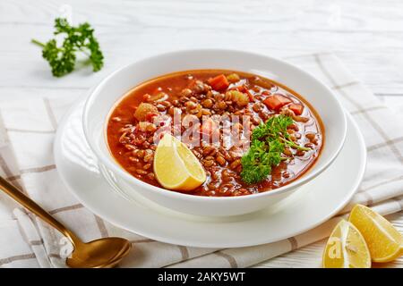 Nahaufnahme von Linsensuppe mit Gemüse und Zitronenkeilen in einer weißen Schüssel auf einem Holztisch mit goldenem Löffel und Toast, horizontale Ansicht von oben Stockfoto