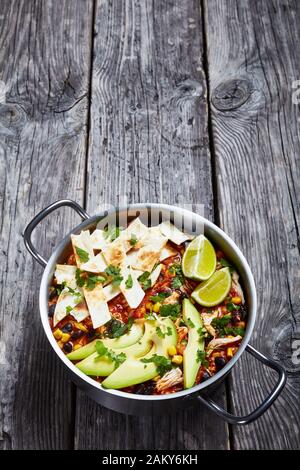 Nahaufnahme von würziger Chicken Taco Suppe mit schwarzer Bohne, Maiskörnchen, mit Tortillastreifen, Avocado-Scheiben und Kalk in einem Metalltopf auf einem rustikalen Woo Stockfoto